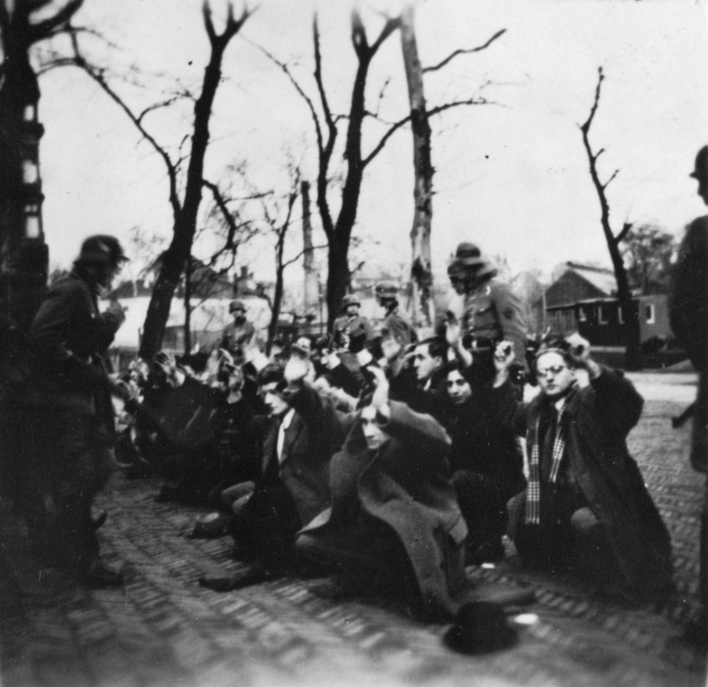 German raid on Jonas Daniël Meijerplein in Amsterdam / Photo of a group of men on their knees, guarded by German soldiers. 25-26 Feb 1941 or 1 May 1940. Photo collection Government Information Service, Photographer unknown/Source National Archive. Public Domain. 