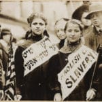 Photograph shows half-length portrait of two girls wearing banners with slogan “ABOLISH CH[ILD] SLAVERY!!” in English and Yiddish (“(ני)דער מיט (קינד)ער שקלאפער(ײ)”, “Nider mit Kinder Schklawerii”), one carrying American flag; spectators stand nearby. Probably taken during May 1, 1909 labor parade in New York City. This work is from the George Grantham Bain collection at the Library of Congress. According to the library, there are no known copyright restrictions on the use of this work.