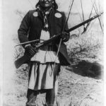 Scene in Geronimo’s camp…before surrender to General Crook, March 27, 1886: Geronimo, full-length portrait standing, facing left, rifle at port, circa 27 March 1886. Photo: C. S. Fly (1849–1901). Public Domain.