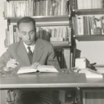 Primo Levi sitting at the desk while reading with a cigarette in his hand – shelves with books in the background, circa 1960. Photo: Anonymous. Public Domain.