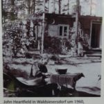 John Heartfield vor seinem Sommerhaus um 1960, Aufnahme auf einer öffentlich zugänglichen Informationstafel vor seinem Sommerhaus in Waldsieversdorf. Photo: Assenmacher. (CC BY-SA 4.0).