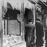 1270px-Germans_walk_by_a_Jewish_business_destroyed_on_Kristallnacht