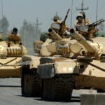 Iraqi tanks assigned to the Iraqi Army 9th Mechanized Division drive through a checkpoint near Forward Operating Base Camp Taji, Iraq. Photo: Taken on 18 May 2006 by U.S. Navy photo by Photographer’s Mate 1st Class Michael Larson (RELEASED). Public Domain.