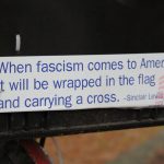 Closeup of bumper sticker with famous Sinclair Lewis line “When fascism comes to America it will be wrapped in the flag and carrying a cross” This quote is often attributed to Sinclair Lewis but has not been verified. The Sinclair Lewis Society notes the following: This quote sounds like something Sinclair Lewis might have said or written, but we’ve never been able to find this exact quote. Date: 27 November 2008. Photo: Robert F. W. Whitlock from Olympia, Washington. (CC BY 2.0).