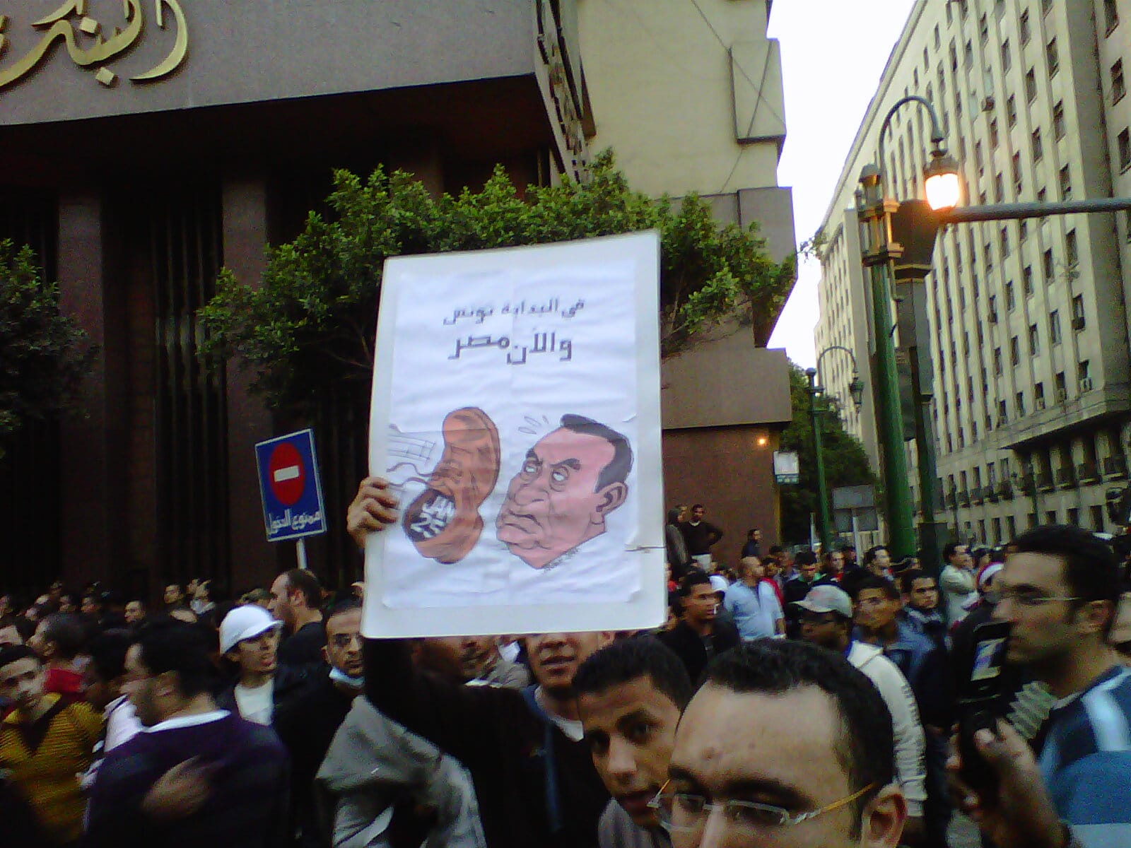 Crowd where a sign with a boot being thrown at Hosni Mubarak is displayed. Author of drawing is Brazilian cartoonist Carlos Latuff. Photo: Taken 25 January 2011 by Muhammad Ghafari from Giza, Egypt. (CC BY 2.0).