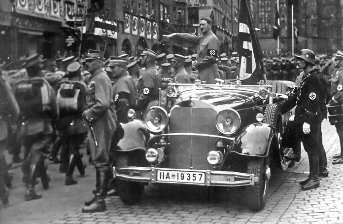 Parade of SA troops past Hitler, Nuremberg 1935. In the car with Hitler: the Blutfahne, behind the car: SS-man en:Jakob Grimminger the carrier of the Blutfahne flag. From the Reichsparteitag der NSDAP 10th-16th September 1935. Author: Charles Russell Collection, NARA. Public Domain. Source: Wikimedia Commons