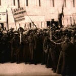 A Soviet revolutionary postcard from the 1917 Russian revolution. Soviet fighters are shown cheering on a street. In the background is a store that used to say in Russian, “Watches, gold and silver.” The has now been altered to read, “Struggle for your rights.” Also a flag that before was a solid color now reads, “Down with the monarchy – long live the Republic!”. Photo: Unknown Soviet photographer. Public Domain.