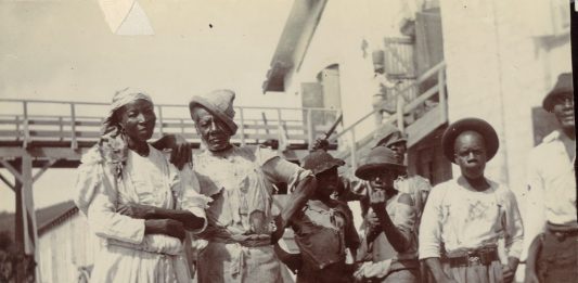 En ældre mand og en kvinde fotograferet sammen med tre drenge og en anden, yngre, mand, på Centralfaktoriets Saftstation, Skt. Croix, 1903. I baggrunden ses en bro, trappeopgang og en bygning. Til venstre yderst i billedet ses et muldyr. Fotograf : Ukendt. Tekst: Henriette Gavnholdt Jacobsen/M/S Museet for Søfart. (CC-BY-NC-SA).