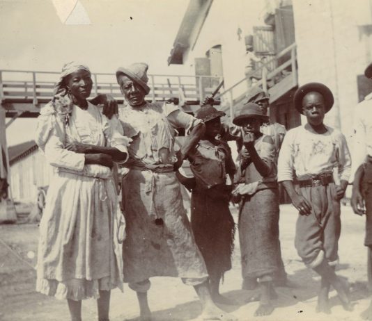 En ældre mand og en kvinde fotograferet sammen med tre drenge og en anden, yngre, mand, på Centralfaktoriets Saftstation, Skt. Croix, 1903. I baggrunden ses en bro, trappeopgang og en bygning. Til venstre yderst i billedet ses et muldyr. Fotograf : Ukendt. Tekst: Henriette Gavnholdt Jacobsen/M/S Museet for Søfart. (CC-BY-NC-SA).